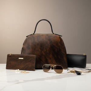 Pode incluir: Uma cole&ccedil;&atilde;o de acess&oacute;rios em couro de vaca castanho e preto, incluindo uma mala, uma carteira e uma pequena bolsa. &Oacute;culos de sol com arma&ccedil;&atilde;o dourada, chaves e um smartphone tamb&eacute;m est&atilde;o presentes. A mala tem uma al&ccedil;a preta.