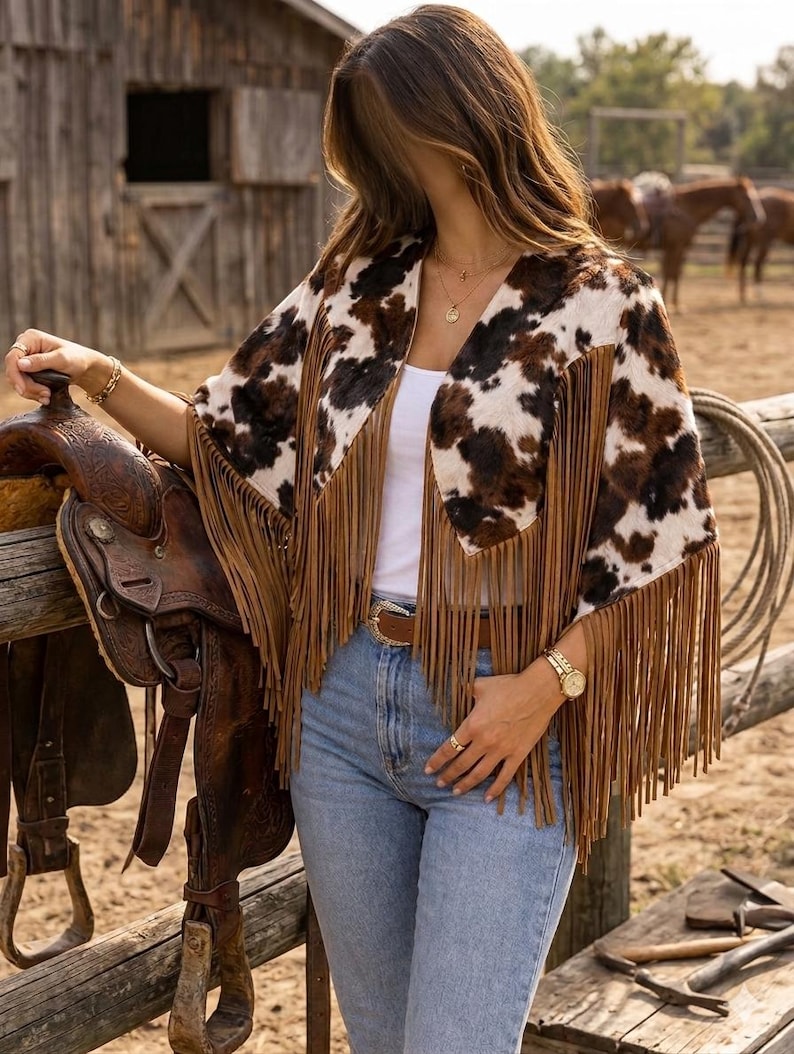 May include: A cow print poncho featuring long brown fringe trim along the edges. The garment is worn over a white top and light blue denim jeans with a brown leather belt. The poncho has a brown and white spotted pattern throughout.