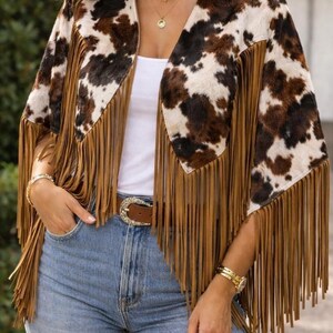 Western faux cowhide fringe cape wrap. Realistic brown and white hair-on-hide pattern with a V-neckline. Features long, tan suede-style tassels along the hem. A unique, boho festival outerwear piece shown styled with light wash jeans and a cowboy buckle belt. Perfect for cowgirl chic, Coastal Cowboy, or rodeo fashion. Handcrafted quality.