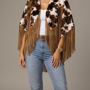 May include: A cow print poncho featuring a brown and white spotted pattern with long tan fringe trim along the edges. The garment is worn over a white tank top and light blue denim jeans with a brown leather belt and brown boots.