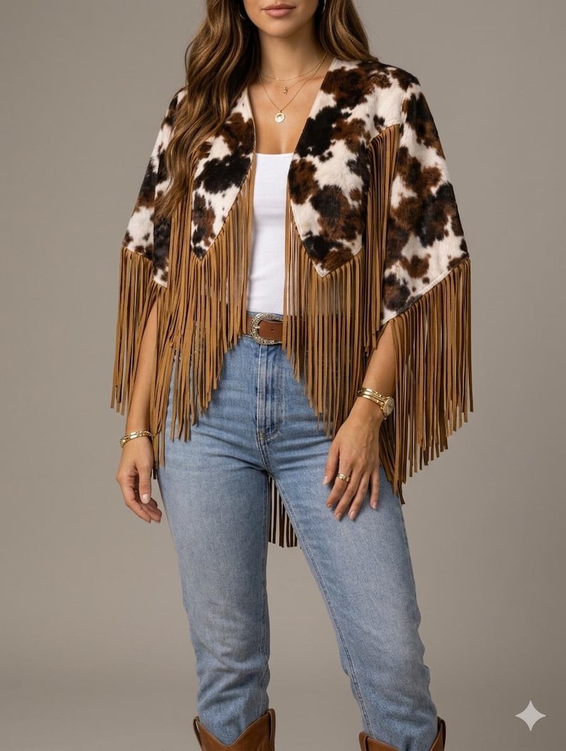 May include: A cow print poncho featuring a brown and white spotted pattern with long tan fringe trim along the edges. The garment is worn over a white tank top and light blue denim jeans with a brown leather belt and brown boots.