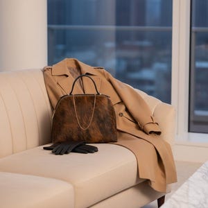 Pode incluir: Um trench coat bege e uma mala com padr&atilde;o castanho e preto com uma al&ccedil;a de corrente dourada repousam num sof&aacute; creme. Luvas de couro pretas s&atilde;o colocadas por baixo da mala. A cena passa-se numa sala bem iluminada com vista para a cidade.