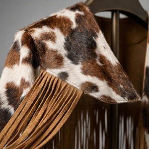 Detail shot of a Western fringe poncho featuring authentic cowhide print and long tan tassels. Close-up view of the high-quality stitching and texture of the faux fur cow print material. Perfect for boho western wear, rodeo outfits, and coastal cowboy fashion enthusiasts looking for high-quality craftsmanship.