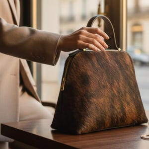 Pode incluir: Uma mala com padr&atilde;o castanho e preto com uma pega superior e fecho de correr dourado. A mala est&aacute; sobre uma mesa de madeira castanha escura. A mala tem uma forma estruturada e parece ser feita de um material texturizado.