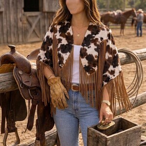 May include: A cow print jacket featuring long brown fringe along the hem and sleeves, worn over a white top and light blue jeans. The jacket has a brown and white spotted pattern, a wide collar, and is paired with a brown leather belt and tan gloves.