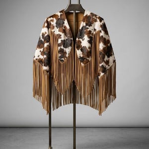 Puede incluir: Chaqueta con estampado de piel de vaca con manchas marrones, negras y blancas. La chaqueta presenta largas y finas flecos color canela en la parte inferior y las mangas. Se exhibe en una percha y soporte de metal, mostrando un diseño de inspiración occidental.