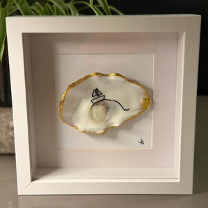 Pode incluir: Uma obra de arte emoldurada com uma concha de ostra branca com uma borda dourada. Um desenho a traço preto de um veleiro está na concha. A obra de arte está em uma moldura branca.