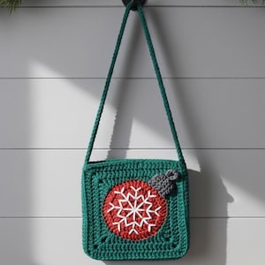 Puede incluir: Un bolso cruzado de ganchillo verde con un círculo rojo y un diseño de copo de nieve blanco. El bolso tiene una correa verde y un pequeño detalle de lazo gris. El bolso está colgado de un gancho negro contra una pared blanca.
