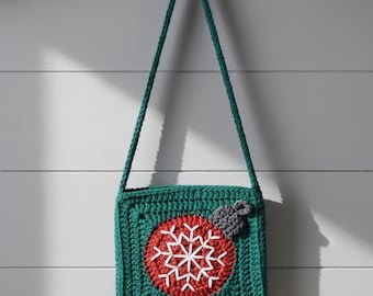 May include: A green crocheted crossbody bag with a red circle and white snowflake design. The bag has a green strap and a small gray bow detail. The bag is hanging on a black hook against a white wall.