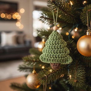 May include: A green crocheted Christmas tree ornament with a brown trunk hangs on a decorated Christmas tree. The tree is adorned with gold ornaments and lights, creating a festive holiday scene. The ornament is suspended by a green string and a wooden bead.