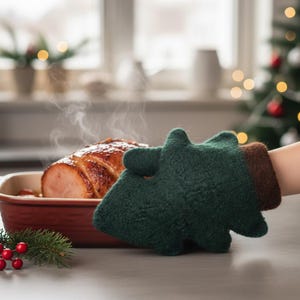 May include: A green felt Christmas tree-shaped oven mitt holds a red baking dish with a cooked ham. The ham is steaming, and a Christmas tree is in the background. A sprig of greenery with red berries is in the foreground.