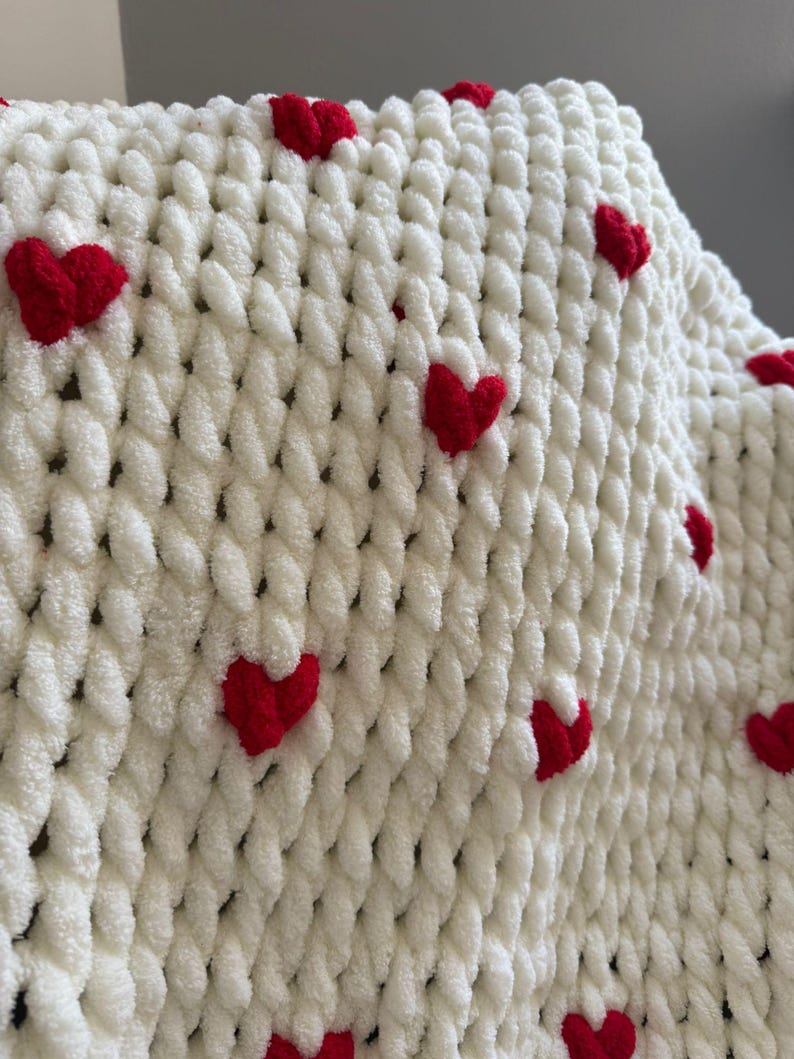 May include: A white, chunky knit blanket adorned with evenly spaced, bright red hearts. The blanket's texture is soft and plush, with the red hearts adding a pop of color and a touch of whimsy. This cozy blanket is perfect for snuggling.