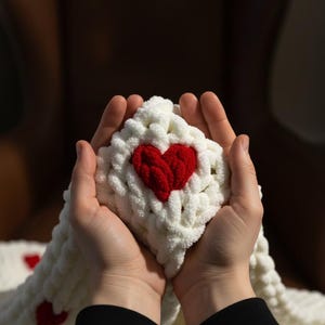 May include: A white, chunky-knit blanket with a large, red heart in the center. The blanket is held in cupped hands, with a black sleeve visible at the bottom. The blanket appears soft and cozy, perfect for Valentine's Day or a gift.