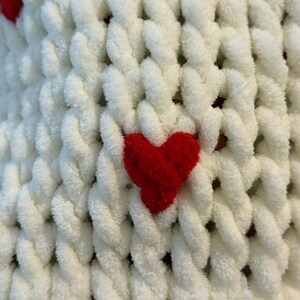 May include: Close-up of a white, chunky knit blanket with red heart accents. The blanket has a textured, looped design, with the red hearts evenly spaced across the surface. The soft, plush material suggests warmth and comfort.