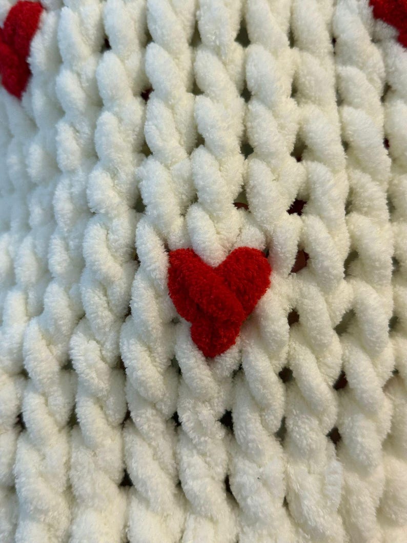 May include: Close-up of a white, chunky knit blanket with red heart accents. The blanket has a textured, looped design, with the red hearts evenly spaced across the surface. The soft, plush material suggests warmth and comfort.