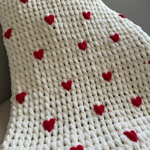 May include: A white, chunky knit blanket with red heart accents. The blanket is made of a soft, textured material and features multiple embroidered red hearts scattered across the surface. The blanket is folded, showing its thick, cozy texture.