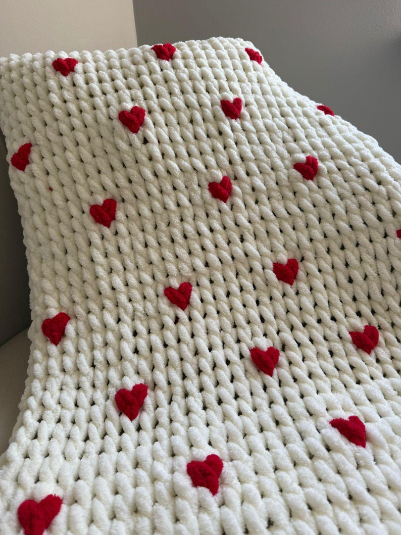 May include: A white, chunky knit blanket with red heart accents. The blanket is made of a soft, textured material and features multiple embroidered red hearts scattered across the surface. The blanket is folded, showing its thick, cozy texture.