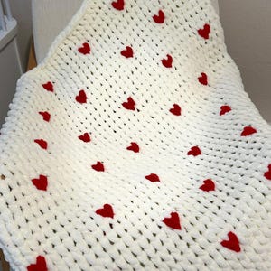 May include: A white, chunky knit blanket with evenly spaced red hearts. The blanket is draped over a chair, showcasing its soft texture and the vibrant contrast of the red hearts against the white background. A cozy, handmade item.
