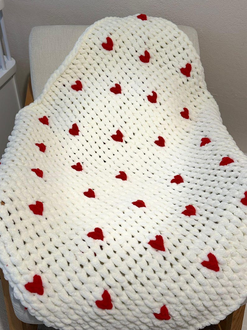 May include: A white, chunky knit blanket with evenly spaced red hearts. The blanket is draped over a chair, showcasing its soft texture and the vibrant contrast of the red hearts against the white background. A cozy, handmade item.