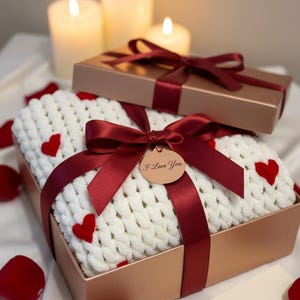 May include: A gift box with a white knitted blanket featuring red heart accents, tied with a burgundy ribbon and a tag that says "I Love You". A smaller box sits on top, also tied with a burgundy ribbon. Lit candles and rose petals complete the romantic scene.