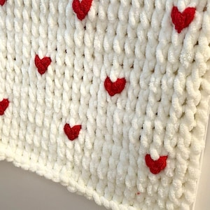 May include: A white, chunky knit blanket with evenly spaced, bright red heart appliqu&eacute;s. The blanket has a soft, plush texture and a thick, braided edge. The hearts are small and add a pop of color to the otherwise neutral design.