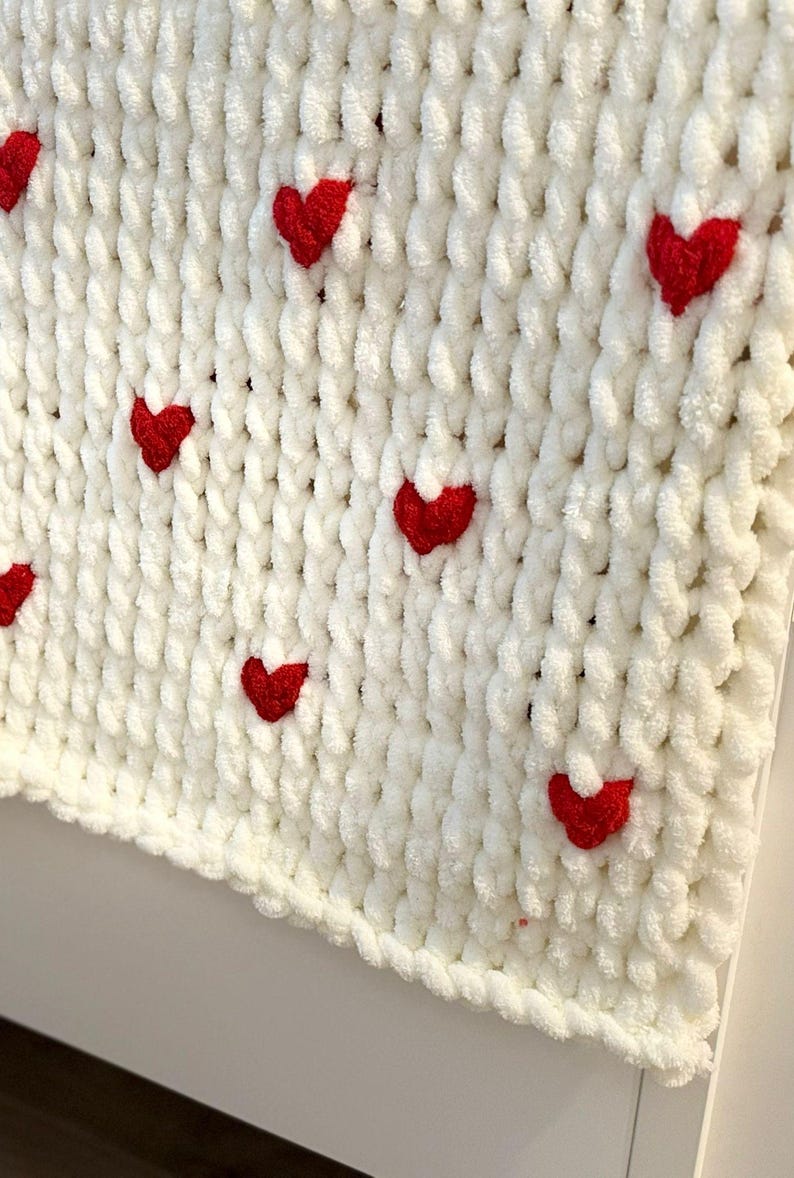 May include: A white, chunky knit blanket with evenly spaced, bright red heart appliqu&eacute;s. The blanket has a soft, plush texture and a thick, braided edge. The hearts are small and add a pop of color to the otherwise neutral design.