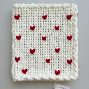 May include: A cream-colored, chunky knit blanket adorned with evenly spaced red heart appliqu&eacute;s. The blanket has a textured appearance and a decorative border. A small tag with the text "Handmade with Love" is visible.