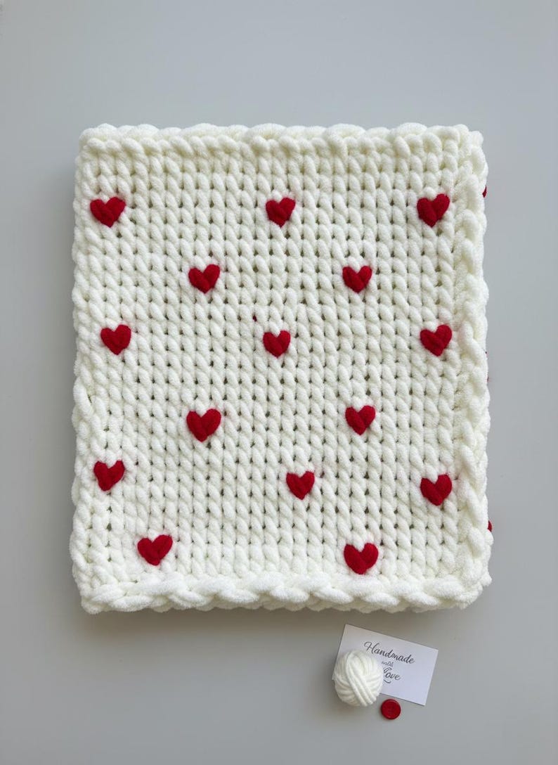 May include: A cream-colored, chunky knit blanket adorned with evenly spaced red heart appliqu&eacute;s. The blanket has a textured appearance and a decorative border. A small tag with the text "Handmade with Love" is visible.