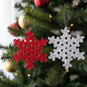 Może przedstawiać: Dwie ręcznie robione ozdoby w kształcie płatków śniegu zrobione na szydełku. Jeden jest w kolorze jasnoczerwonym, drugi biały. Oba wiszą na srebrnych sznurkach na choince, z innymi ozdobami i światłami w tle. Te świąteczne dekoracje.