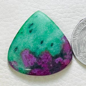 Könnte beinhalten: Ein polierter, dreieckiger Edelstein mit einem leuchtend grünen Oberteil und einer Basis aus tiefem Lila und Schwarz. Der Stein befindet sich neben einer US-Vierteldollarmünze, die als Größenreferenz dient. Das einzigartige Farbmuster des Edelsteins macht ihn zu einem Blickfang.
