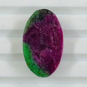 May include: An oval gemstone displaying a striking colour gradient. The stone transitions from a deep magenta in the centre to a green hue on one edge, with a touch of black at the top. The polished surface reflects light.