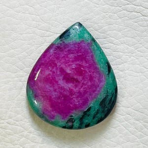 Może przedstawiać: Kształtny kamień szlachetny w kształcie łzy z żywym, purpurowym środkiem i zieloną obwódką. Polerowana powierzchnia ukazuje naturalne wariacje kolorów i wzory. Kamień szlachetny jest umieszczony na białym tle.