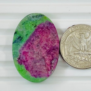 May include: An oval-shaped gemstone with a vibrant mix of colors, including green and a deep magenta. Next to the gemstone is a United States quarter coin, providing a size reference. The gemstone's surface has a natural, textured appearance.
