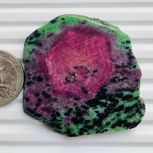 Könnte beinhalten: Ein grob geschnittener Edelstein mit einem lebendigen Farbmuster. Der Stein hat einen zentralen Bereich in tiefem Magenta, umgeben von Grün und mit schwarzen Flecken versehen. Eine Vierteldollarmünze liegt daneben zum Größenvergleich.