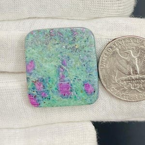 Puede incluir: Una piedra preciosa de forma cuadrada con una mezcla de tonos verdes y rosas. Una moneda de veinticinco centavos estadounidense está al lado para comparar el tamaño. La superficie de la piedra preciosa tiene un aspecto moteado, con zonas rosas en racimos.