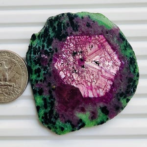 May include: A polished gemstone slice with a striking colour palette. The centre features a pink and white crystalline pattern, surrounded by a deep purple hue. The outer edge is a speckled green and black. A US quarter coin is next to the gemstone.