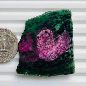 May include: A raw, polished ruby in zoisite gemstone. The stone displays a vibrant mix of deep green and black zoisite with a central cluster of rich, pinkish-red ruby. A US quarter is next to the stone for size comparison.