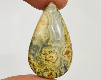 May include: A teardrop-shaped gemstone with a polished surface. The stone features a mix of colors, including beige, yellow, and green, with darker spots and patterns. The gemstone is held between two fingers.
