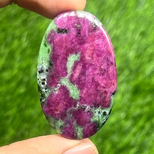May include: An oval-shaped gemstone with a vibrant mix of colors. The stone features a deep magenta hue, interspersed with patches of light green and black speckles. The polished surface reflects light, highlighting the natural patterns.
