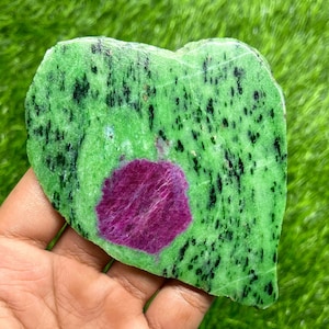 May include: A heart-shaped piece of zoisite with a central ruby inclusion. The zoisite is a vibrant green with black speckles, and the ruby is a deep magenta color. The stone is held in a hand.