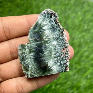 May include: A polished slab of dark green seraphinite mineral featuring a distinct feathery, chatoyant pattern with radiating silver-white inclusions and subtle reddish-brown edges, displaying a smooth, reflective surface texture throughout.
