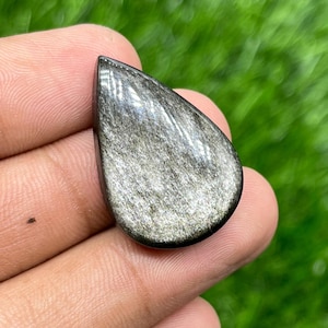 Puede incluir: Una gema pulida en forma de lágrima con un brillo plateado metálico. La piedra tiene un color base oscuro, casi negro, con inclusiones plateadas brillantes, creando una superficie reflectante. La gema se sostiene en una mano.