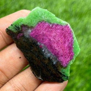 May include: A rough-cut gemstone with a vibrant color palette. The stone features a central magenta section, bordered by a green layer, and a dark, almost black base. The textures vary from rough to smooth.