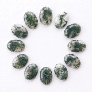 Pu&ograve; includere: Una collezione di undici pietre preziose di agata muschiata di forma ovale disposte in un motivo circolare. Ogni pietra lucidata presenta una base bianca traslucida con distinte inclusioni dendritiche verde scuro simili a felci.