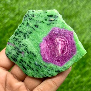 May include: A rough-cut ruby zoisite stone. The stone features a vibrant green base with black speckles and a central, hexagonal-shaped ruby in shades of pink and purple. The stone is irregularly shaped.