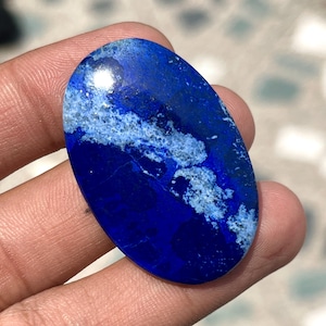 May include: Oval-shaped lapis lazuli gemstone. The stone features a deep blue color with white and gray marbling. The polished surface reflects light, highlighting the stone's natural patterns. This gemstone is suitable for jewelry making.