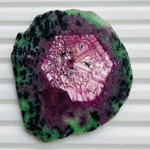 Puede incluir: Una rodaja de piedra preciosa pulida de forma irregular. La piedra presenta una vibrante paleta de colores, con una zona central rosa y blanca, rodeada por una banda morada oscura y bordeada con motas verdes y negras. La superficie de la piedra es lisa y reflectante.