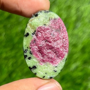 May include: An oval gemstone featuring a central area of deep pink-red, encircled by a light green. The green section is speckled with small black spots. The stone is held between two fingers against a blurred green backdrop.