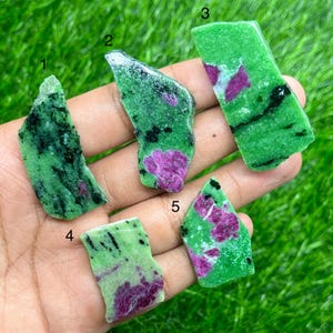 Puede incluir: Cinco piedras de zoisita en bruto con inclusiones de rubí. Las piedras son de un verde vibrante con vetas negras y parches de color rojo-rosado intenso. Las piedras se sostienen en una mano sobre un fondo de hierba verde.