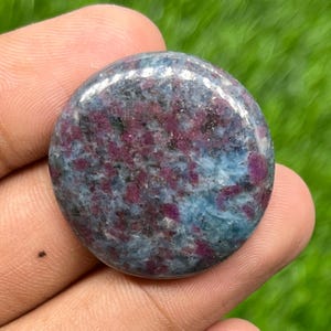 Op de afbeelding: Een gepolijste, ronde edelsteen met een mix van kleuren. De steen heeft een mix van dieprode, blauwe en grijze tinten, waardoor een gemarmerd effect ontstaat. Het oppervlak is glad en reflecterend, wat wijst op een hoogwaardige afwerking. De edelsteen wordt in een hand gehouden.